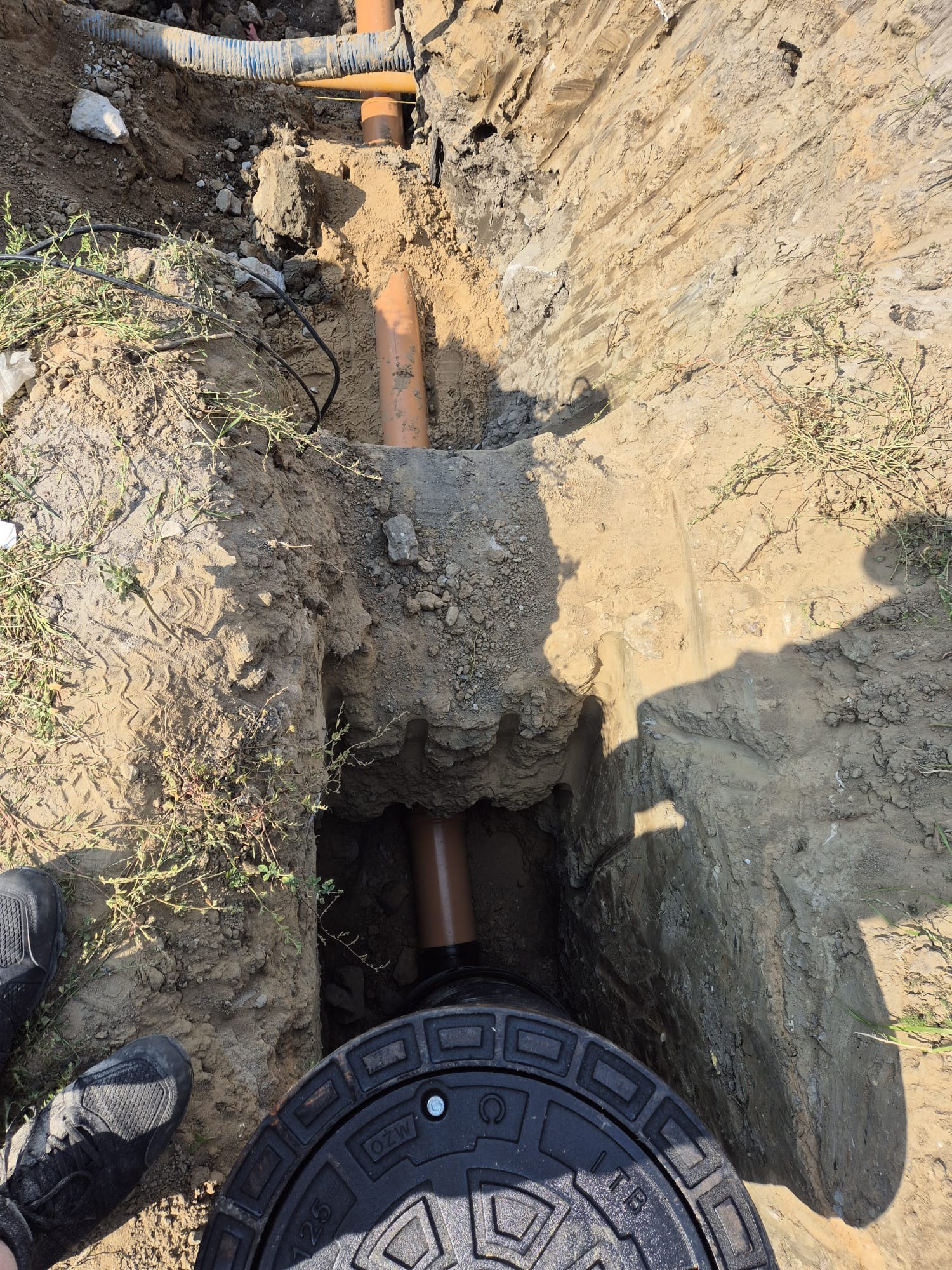 Wykop z widocznymi rurami kanalizacyjnymi z tworzywa sztucznego i czarną pokrywą studzienki rewizyjnej, obok but roboczy, w tle ziemia i trawa.