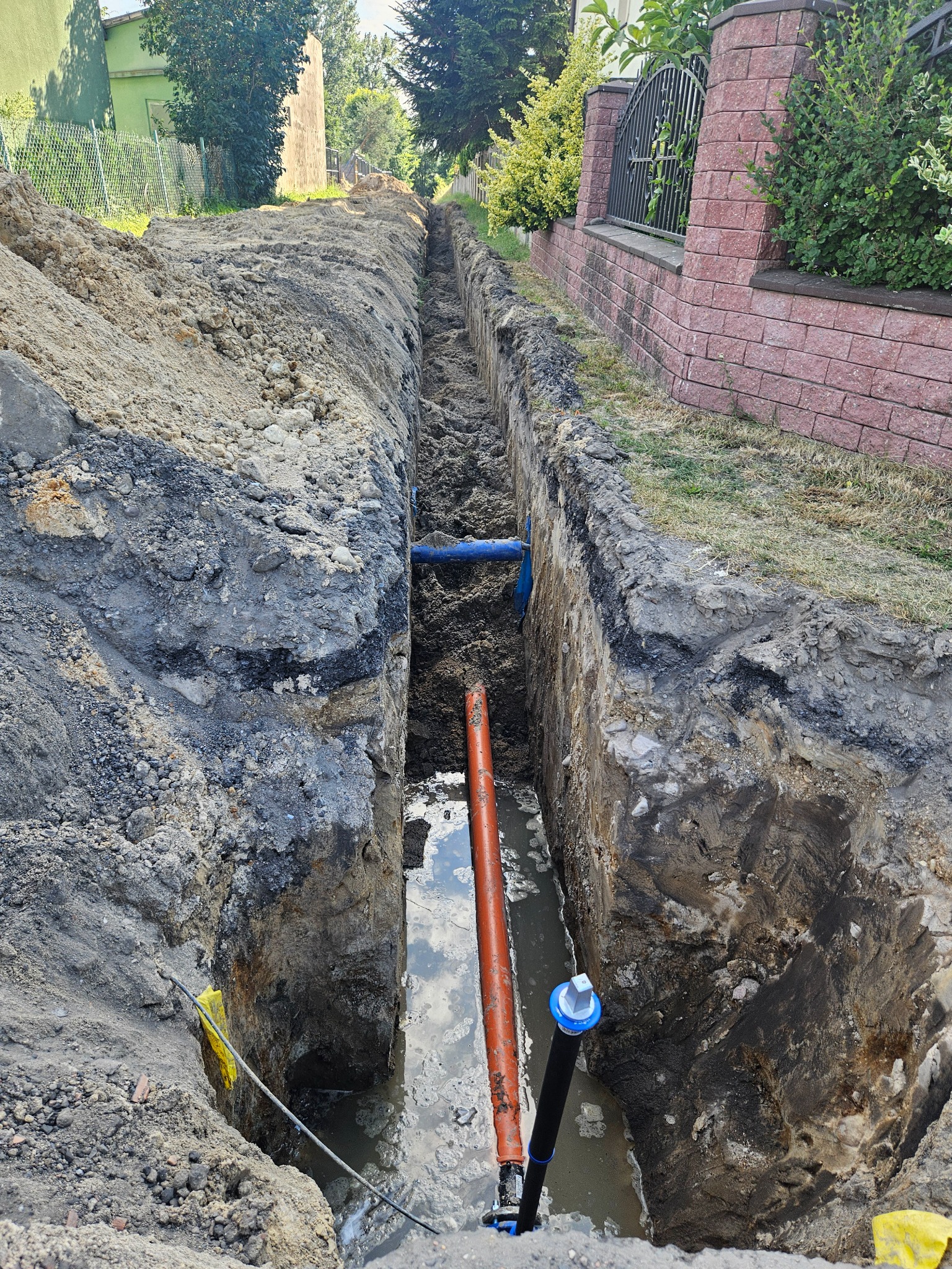 Wykop w ziemi z widocznymi rurami (pomarańczowa i niebieska) częściowo zanurzonymi w wodzie, teren budowy ogrodzony ceglanym murem.