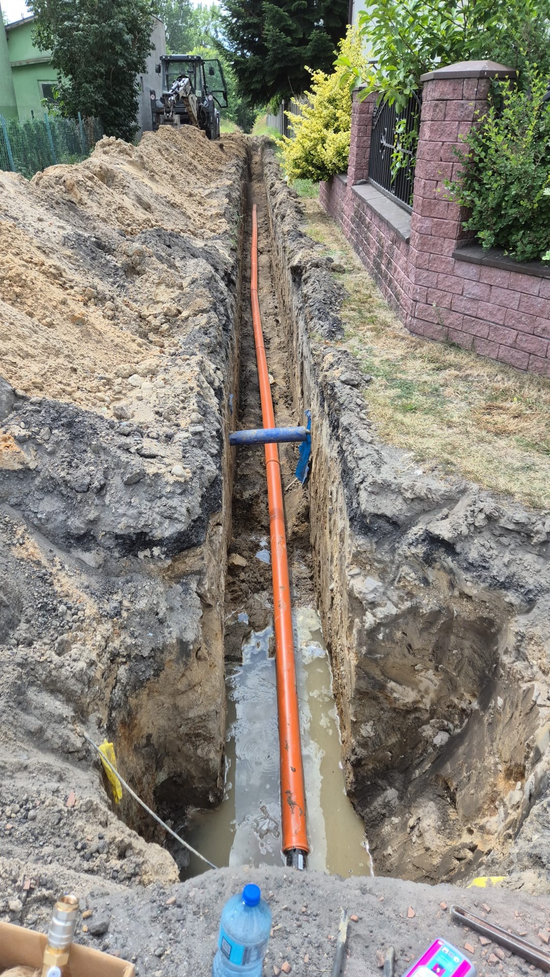 Pomarańczowa rura ułożona w wykopie ziemnym, częściowo wypełnionym wodą, z widocznym niebieskim łącznikiem i koparką w tle.