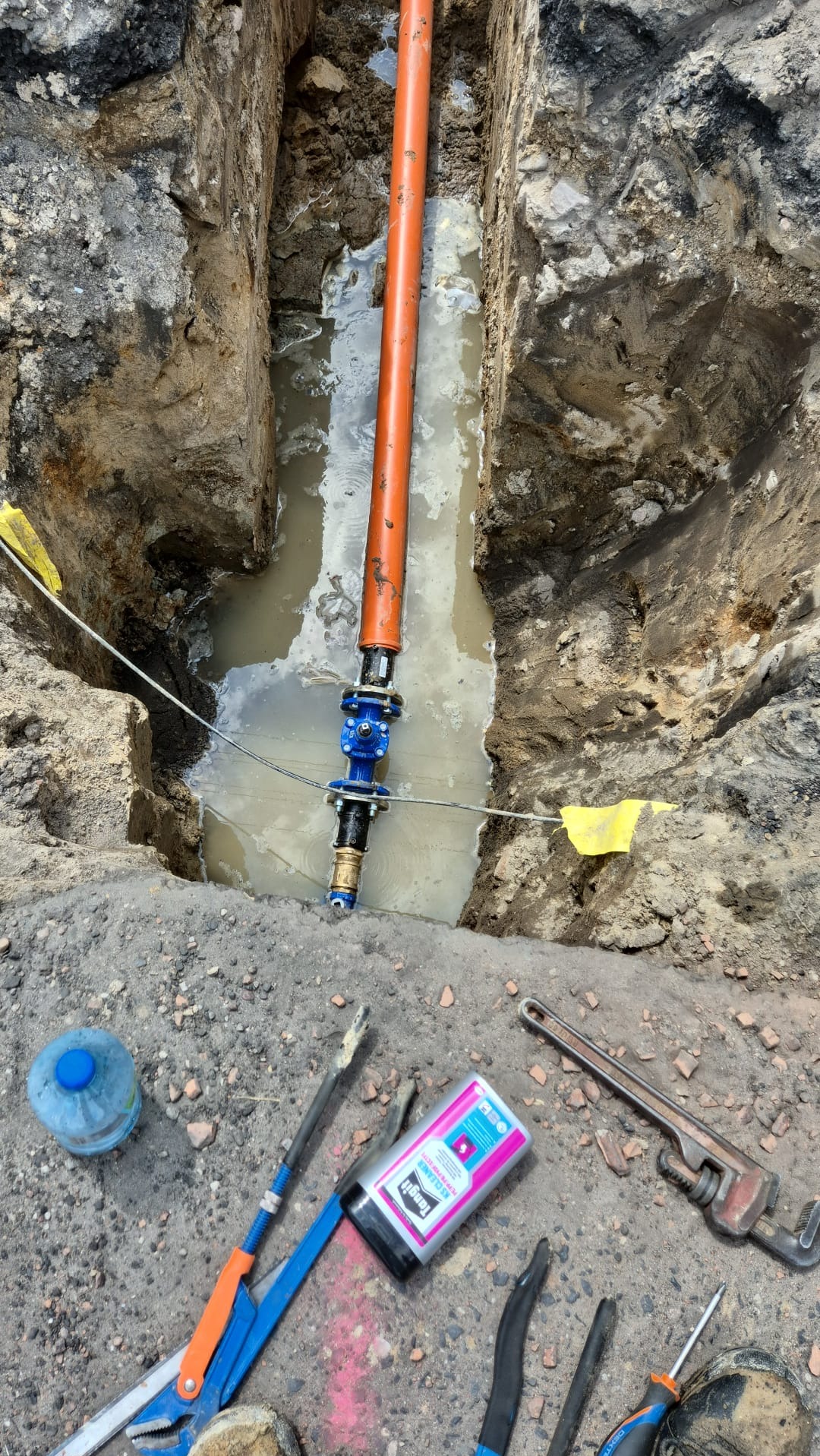 Wykop z wodą, pomarańczowa rura pionowa z niebieskim zaworem, narzędzia hydrauliczne i butelka wody na ziemi.