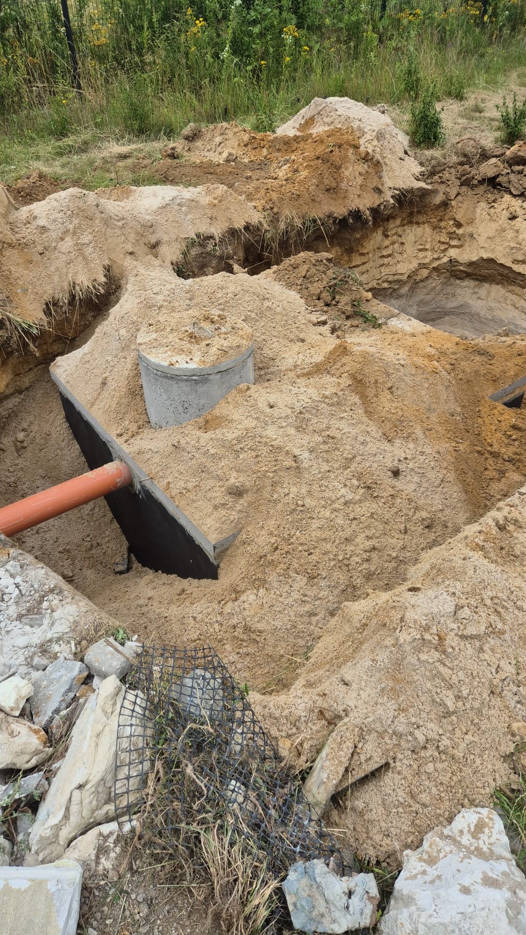 Wykonany wykop pod szambo betonowe, widoczna rura kanalizacyjna, betonowy krąg i piasek.