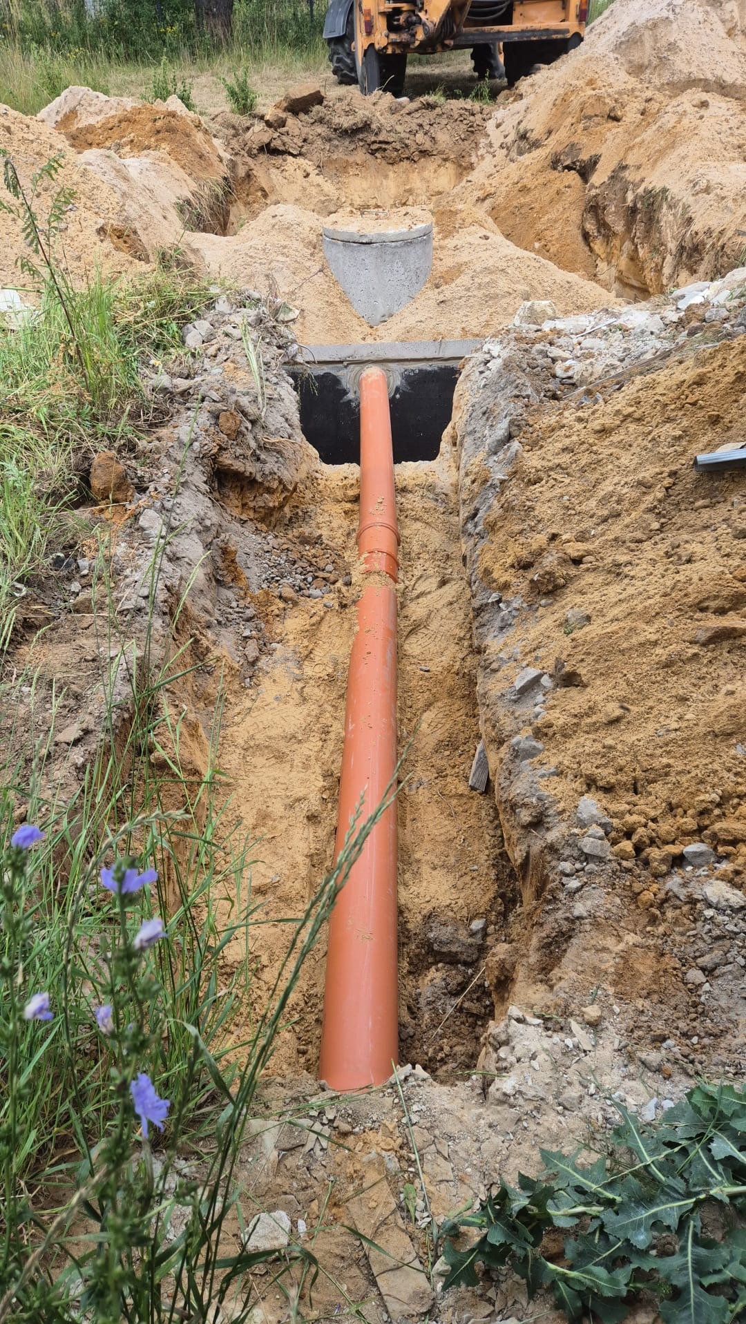 Pomarańczowa rura kanalizacyjna w wykopie z piasku, widoczny betonowy element studzienki i traktor w tle.