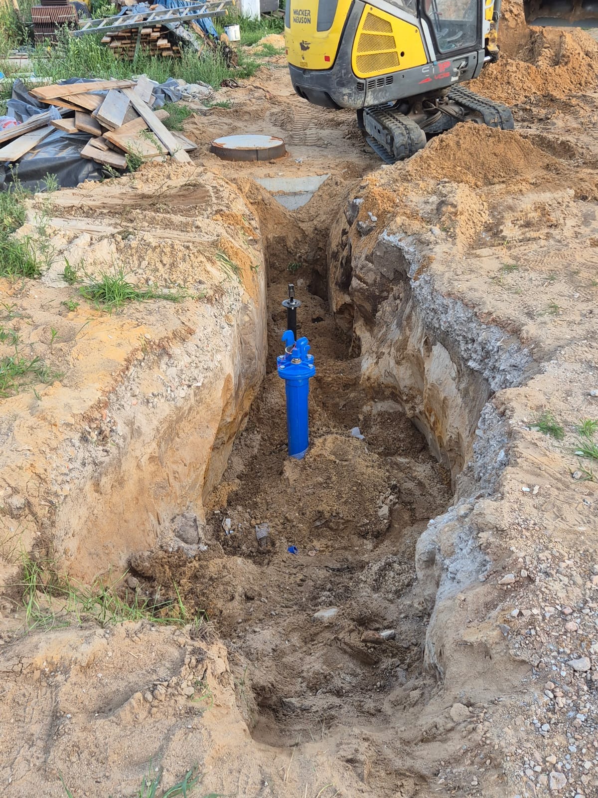 Głęboki wykop w ziemi z zainstalowaną niebieską rurą wodociągową i czarnym zaworem, widoczny właz studzienki i koparka Wacker Neuson w tle.