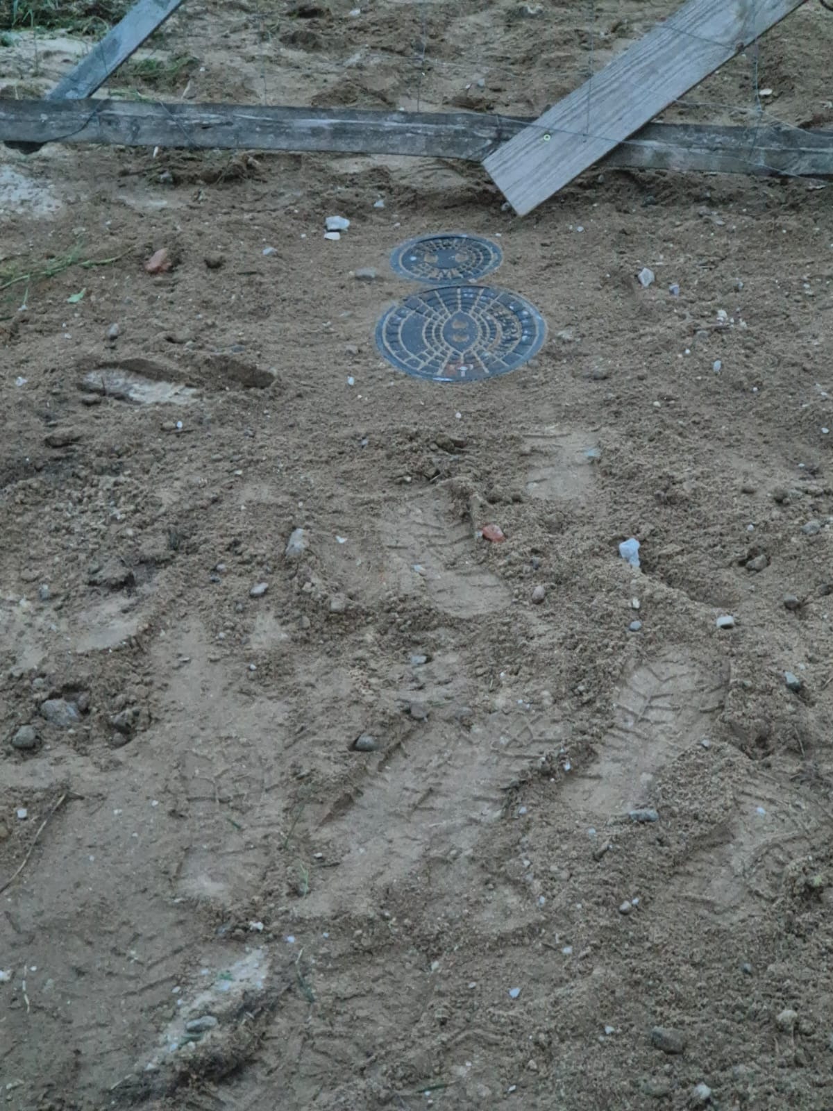 Dwa włazy kanalizacyjne o okrągłym kształcie, jeden większy i jeden mniejszy, wystające z ziemi na placu budowy, otoczone piaskiem i śladami opon, widoczne elementy drewniane konstrukcji w tle.