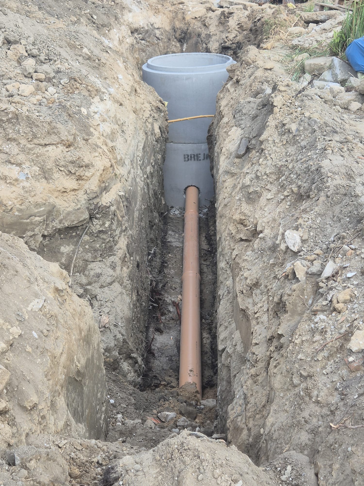 Betonowy zbiornik, prawdopodobnie szambo, częściowo zagłębiony w wykopie, z podłączoną rurą kanalizacyjną. Widoczne ślady świeżych prac ziemnych.