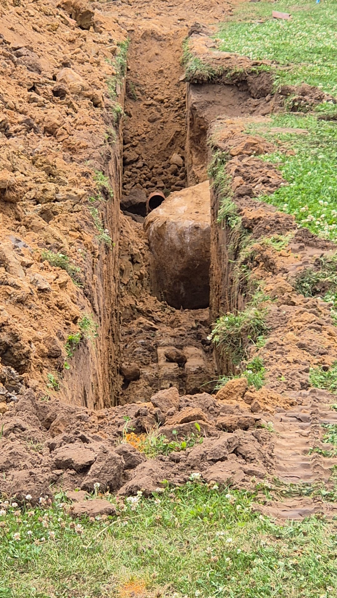 Wykop w ziemi z widocznym fragmentem rury i dużym kamieniem, prawdopodobnie podczas prac ziemnych związanych z instalacją sanitarną.