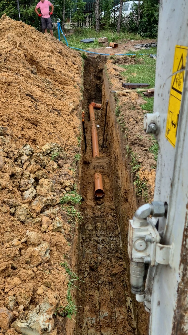 Wykop z ułożonymi rurami kanalizacyjnymi z tworzywa sztucznego, widok z góry na dno wykopu, częściowo zasypane ziemią, z boku widoczny fragment maszyny budowlanej.