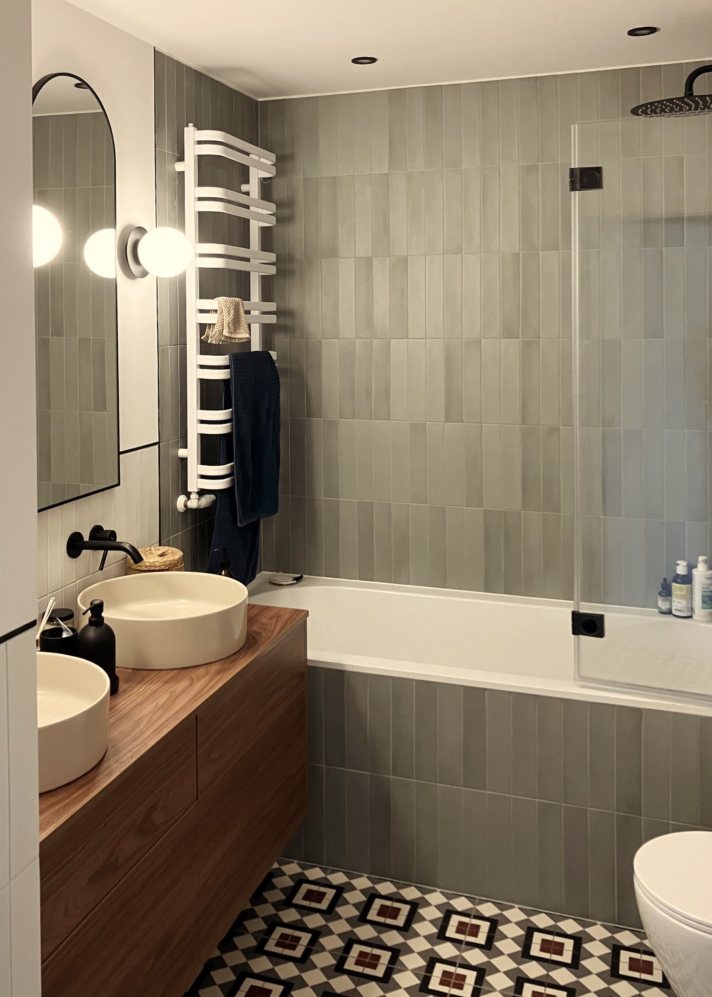 Nowoczesna łazienka z dwoma umywalkami na drewnianym blacie, szare płytki na ścianach i podłodze w geometryczny wzór. Grzejnik drabinkowy i czarna armatura.