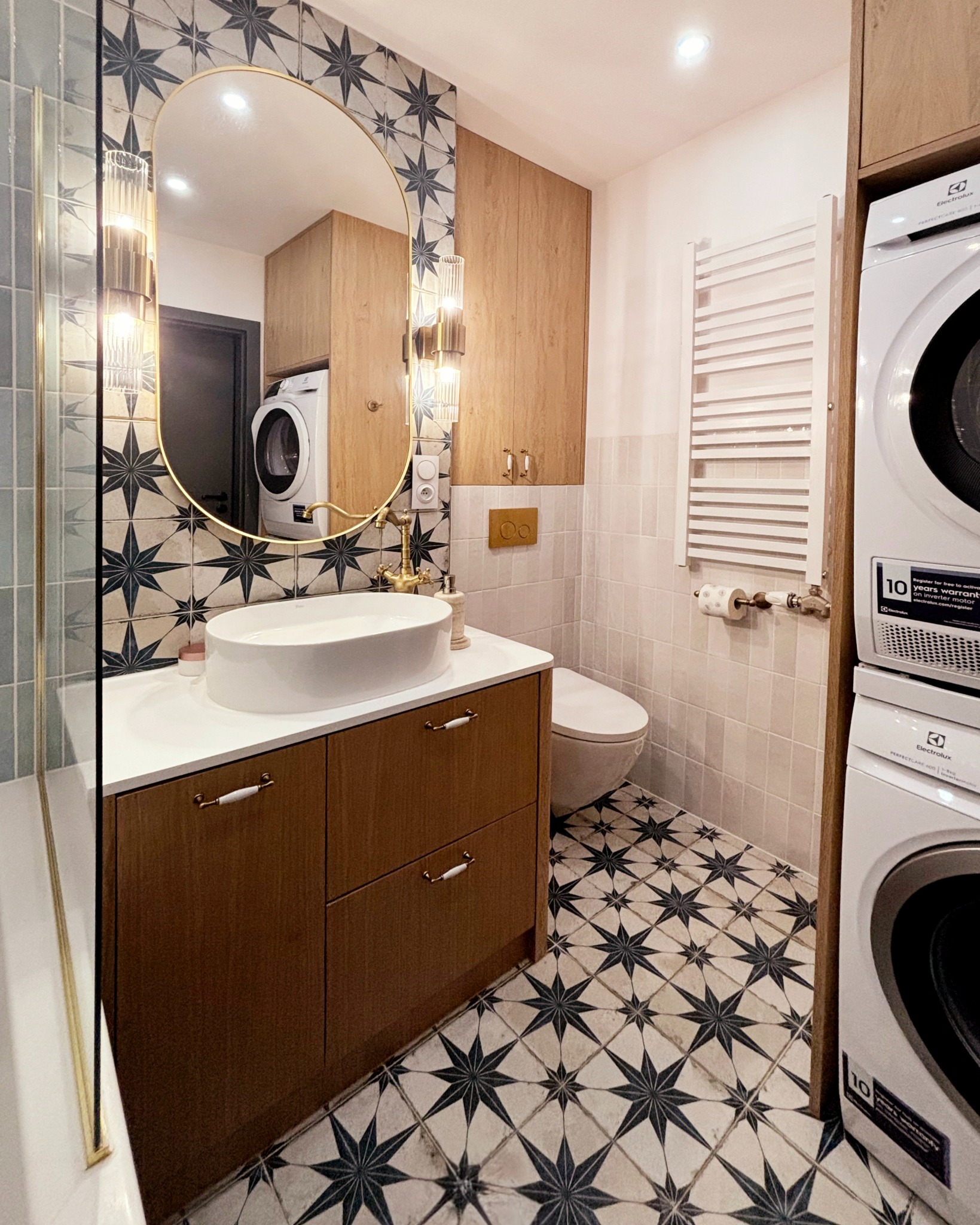 Stylowa łazienka z owalnym lustrem w złotej ramie, wzorzystymi płytkami i drewnianymi szafkami. Widoczna pralka i suszarka Electrolux. Umywalka wpuszczana.