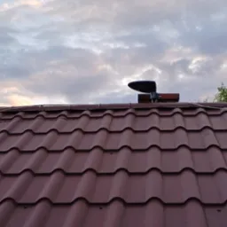 Antena satelitarna zamontowana na brązowym dachu z blachodachówki, na tle pochmurnego nieba o zmierzchu.