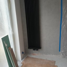 Grzegorz Małecki - Czarny, pionowy grzejnik żeberkowy zamontowany na szarej ścianie w trakcie remontu, z widocznymi płytami gipsowo-kartonowymi i folią ochronną na oknie.