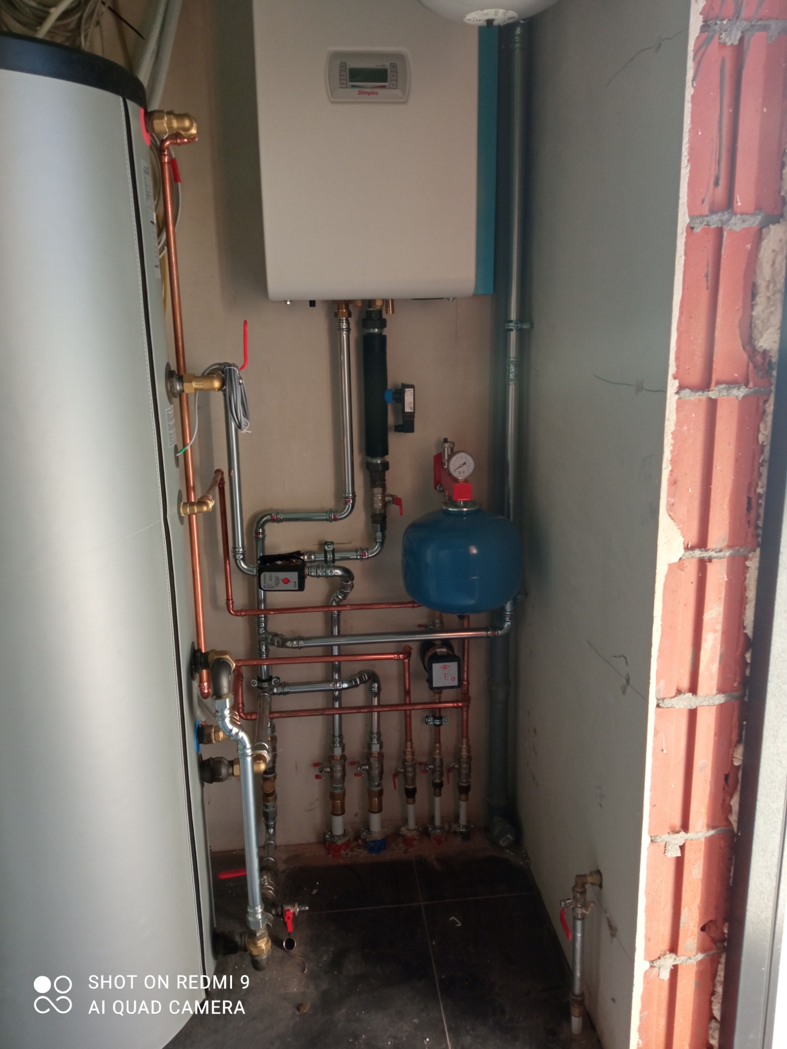 Kompleksowa instalacja grzewcza z piecem gazowym, zbiornikiem wyrównawczym i rurami miedzianymi, zrealizowana w Środzie Wielkopolskiej. Widoczna surowa ściana z cegły obok.