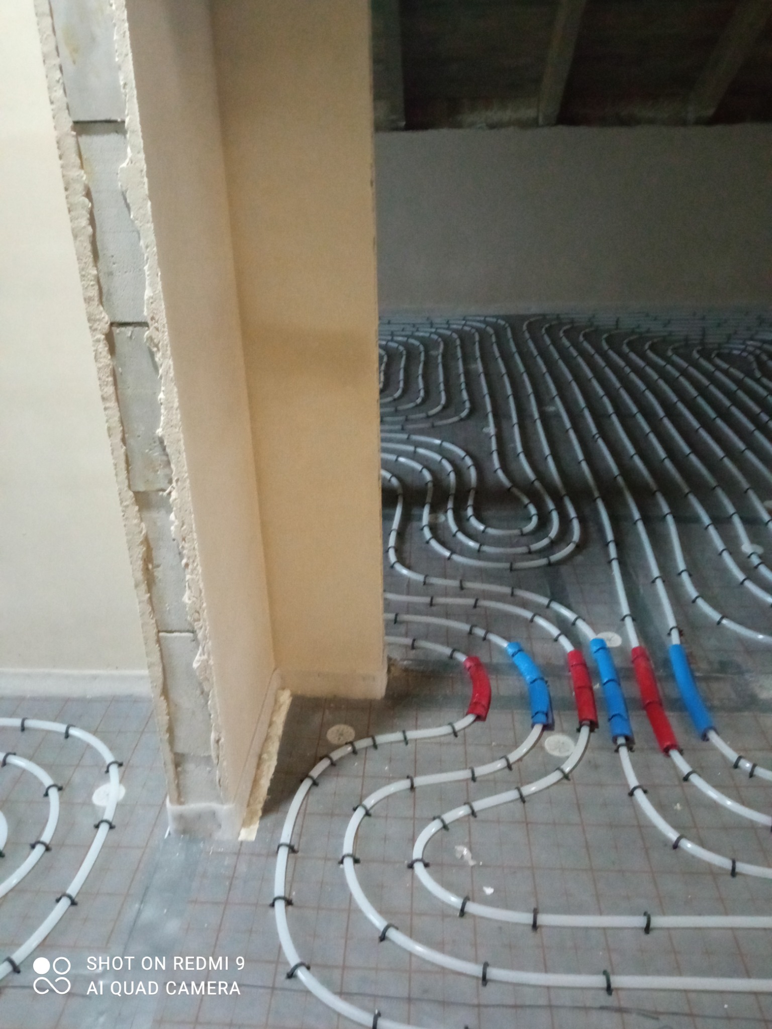 Rury ogrzewania podłogowego, białe z czarnymi mocowaniami, układane w pętle na szarej posadzce, z czerwonymi i niebieskimi oznaczeniami przyłączeń, widok na surowy stan ścian.