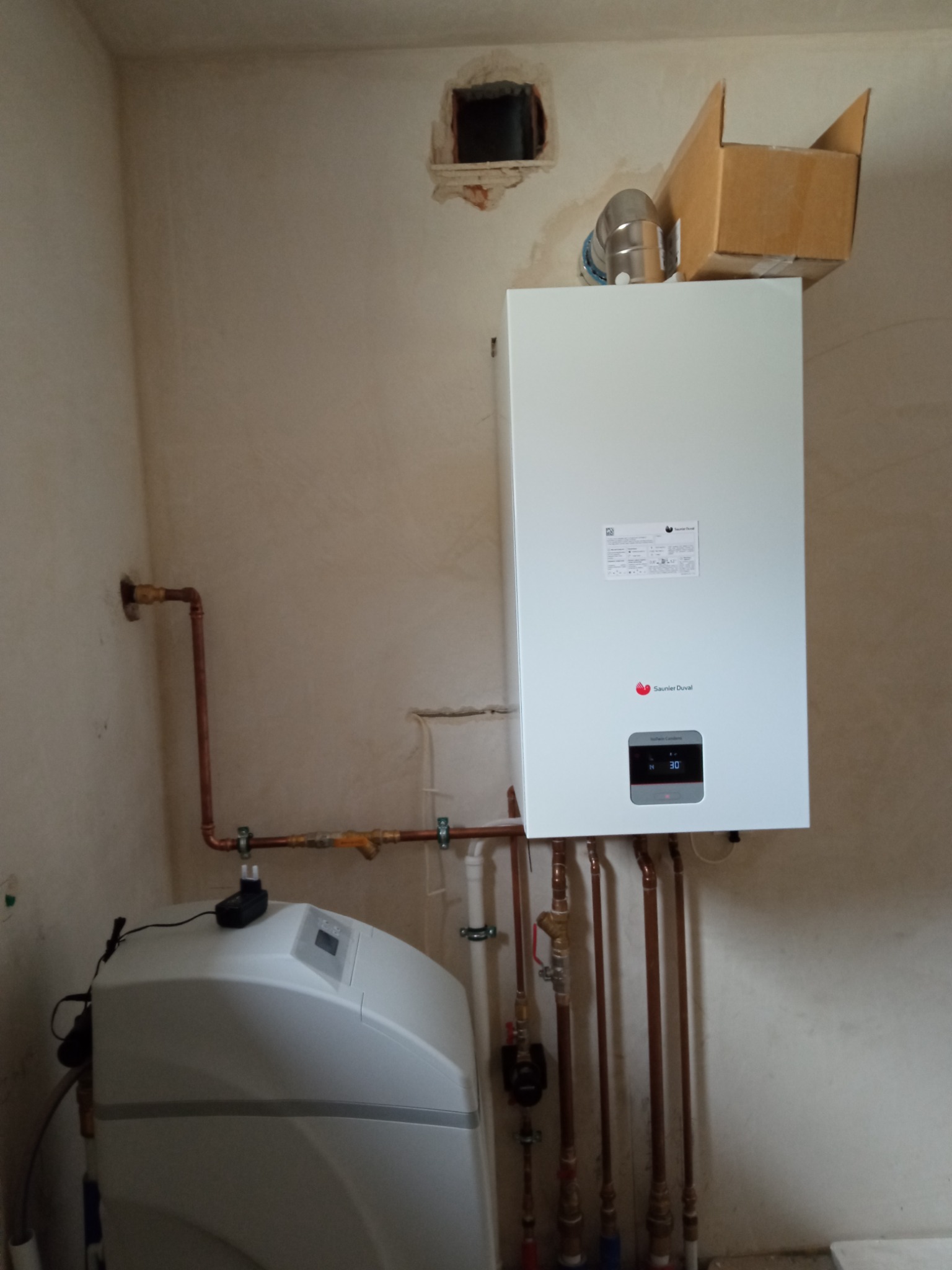 Biały, wiszący piec gazowy marki Saunier Duval zamontowany na ścianie z widocznymi rurami miedzianymi, obok zmiękczacz wody. Na górze pieca widoczny karton i komin spalinowy. Ściana w tle jest...
