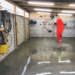 Pracownik w kombinezonie czyszczący posadzkę w garażu za pomocą lancy ciśnieniowej; na ścianach wiszą narzędzia.