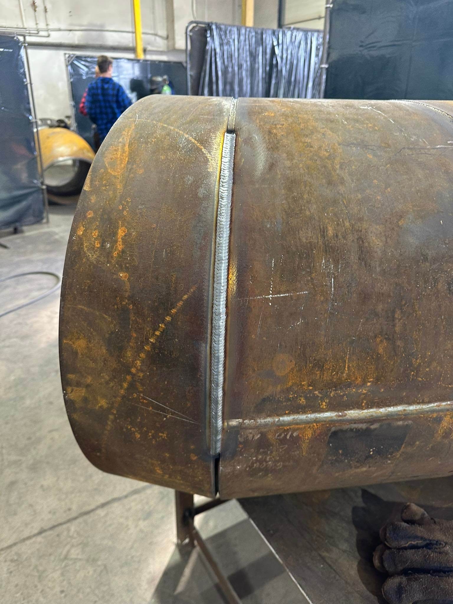 Zespawany pionowo szew na rdzewiejącym, cylindrycznym zbiorniku, widoczne ślady po spawaniu i zarysowania na metalu, w tle postać w niebieskiej koszuli.