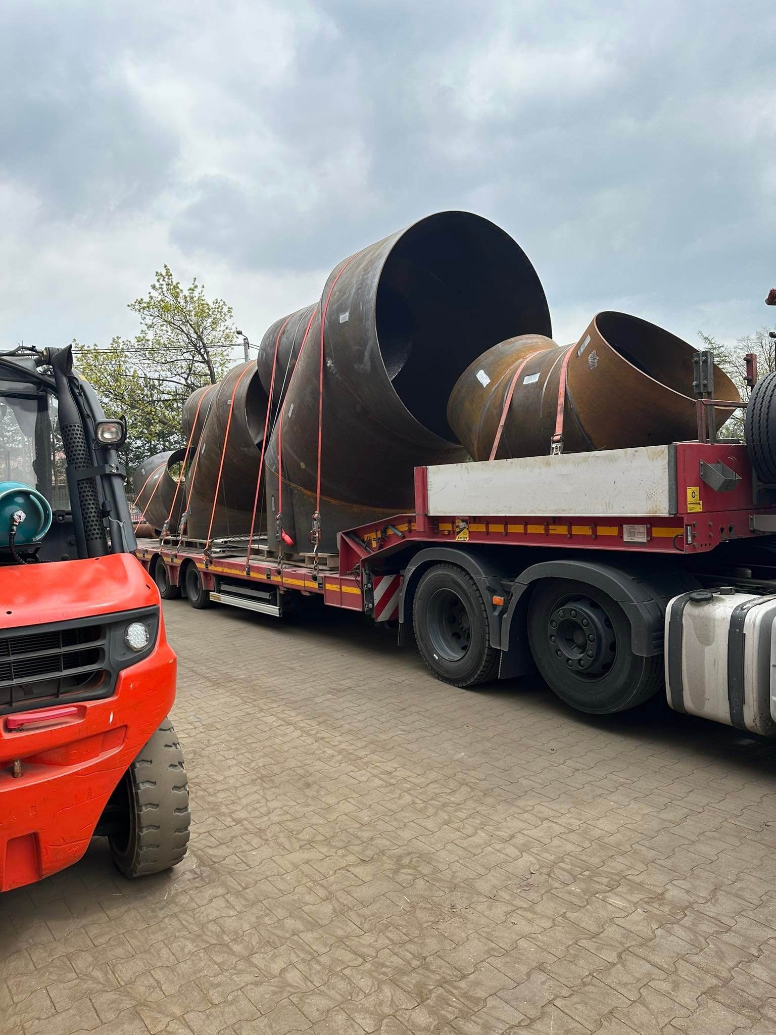 Ciężarówka z naczepą przewozi elementy rurociągu o dużych średnicach, zabezpieczone pasami transportowymi. Widoczny wózek widłowy z butlą gazową w pobliżu.