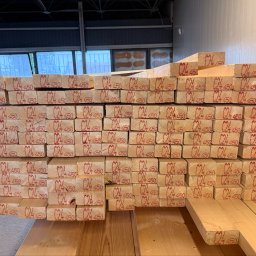 Scandura Homes - Stos jasnych, drewnianych belek konstrukcyjnych z czerwonym stemplem C24 450 na każdym elemencie, ułożonych w magazynie z widocznymi oknami w tle.
