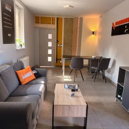 Scandura Homes - Wnętrze showroomu z szarą sofą i stolikiem kawowym. Fragment ściany eksponuje konstrukcję szkieletową z drewnianymi belkami i wełną mineralną, obok plakat 'TECHNOLOGIA SZKIELETOWA'.