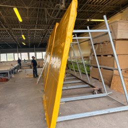 Scandura Homes - Produkcja prefabrykowanych paneli ściennych lub dachowych, owiniętych żółtą folią, opartych o metalowy stojak w hali produkcyjnej. W tle pracownicy i stosy płyt drewnopochodnych.