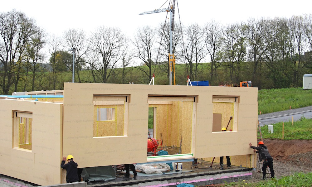 Montaż prefabrykowanej ściany z drewna na budowie domu szkieletowego. Widoczny dźwig i pracownicy w kaskach. Ściana z otworami okiennymi.