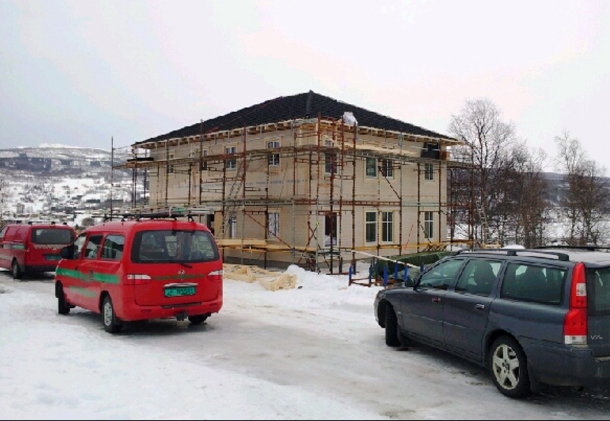 Dwukondygnacyjny dom szkieletowy w trakcie budowy, otoczony rusztowaniami, na tle zimowego krajobrazu. Na pierwszym planie zaparkowane busy i samochód osobowy.