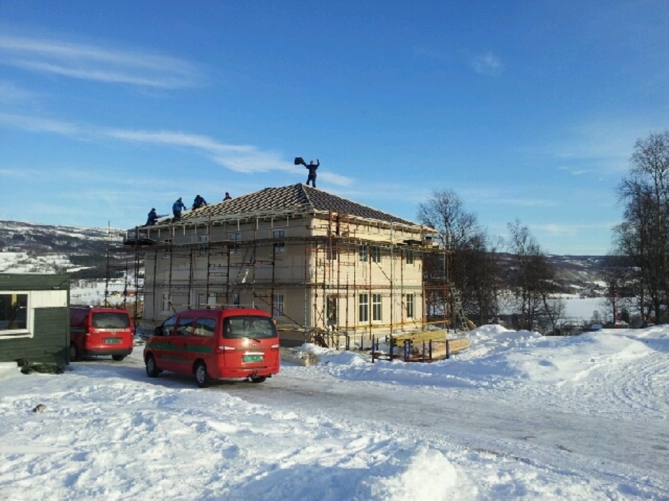 Budowa domu szkieletowego w zimowym krajobrazie Norwegii. Prefabrykowana konstrukcja timber frame, rusztowania, ekipa na dachu. Dwa czerwone busy na śniegu.