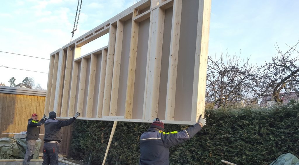 Montaż ściany szkieletowej domu z pomocą dźwigu. Trzech robotników w roboczych ubraniach stabilizuje element. Widoczne drzewa w tle i fragmenty konstrukcji.