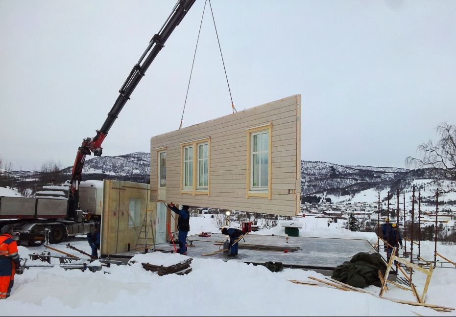 Montaż ściany z oknami domu szkieletowego za pomocą dźwigu na tle zimowego krajobrazu. Widoczni pracownicy w trakcie pracy.
