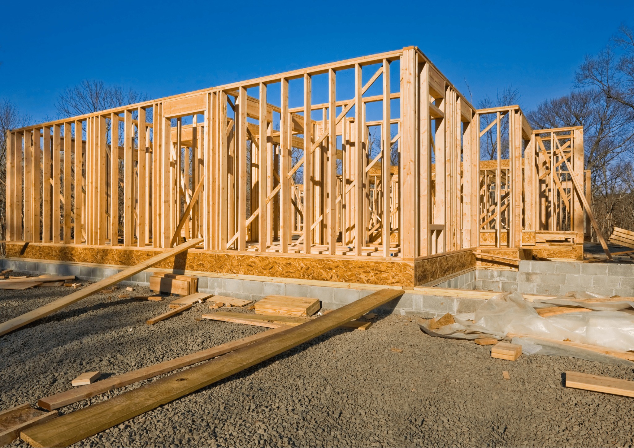 Wznoszenie konstrukcji domu szkieletowego na fundamencie. Widoczne drewniane elementy szkieletu i podłoże z gruzu. Błękitne niebo w tle.