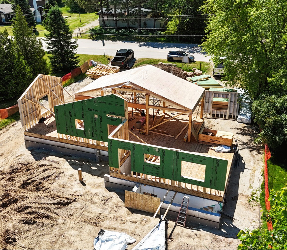 Wysoka perspektywa na budowę domu szkieletowego z zielonymi panelami ściennymi i drewnianą konstrukcją dachu. Widoczne fundamenty i otoczenie z drzewami i drogą.