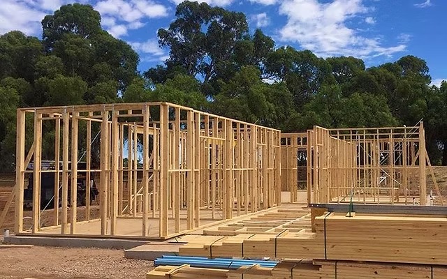 Konstrukcja domu szkieletowego w trakcie budowy, widoczne drewniane ramy ścian i stropu na fundamencie, z drzewami w tle i materiałami budowlanymi na pierwszym planie.