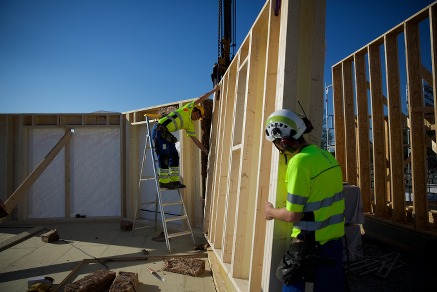 Montaż ściany szkieletowej domu przez dwóch pracowników w odblaskowych ubraniach. Widoczny dźwig i elementy konstrukcyjne, jasne niebo w tle.