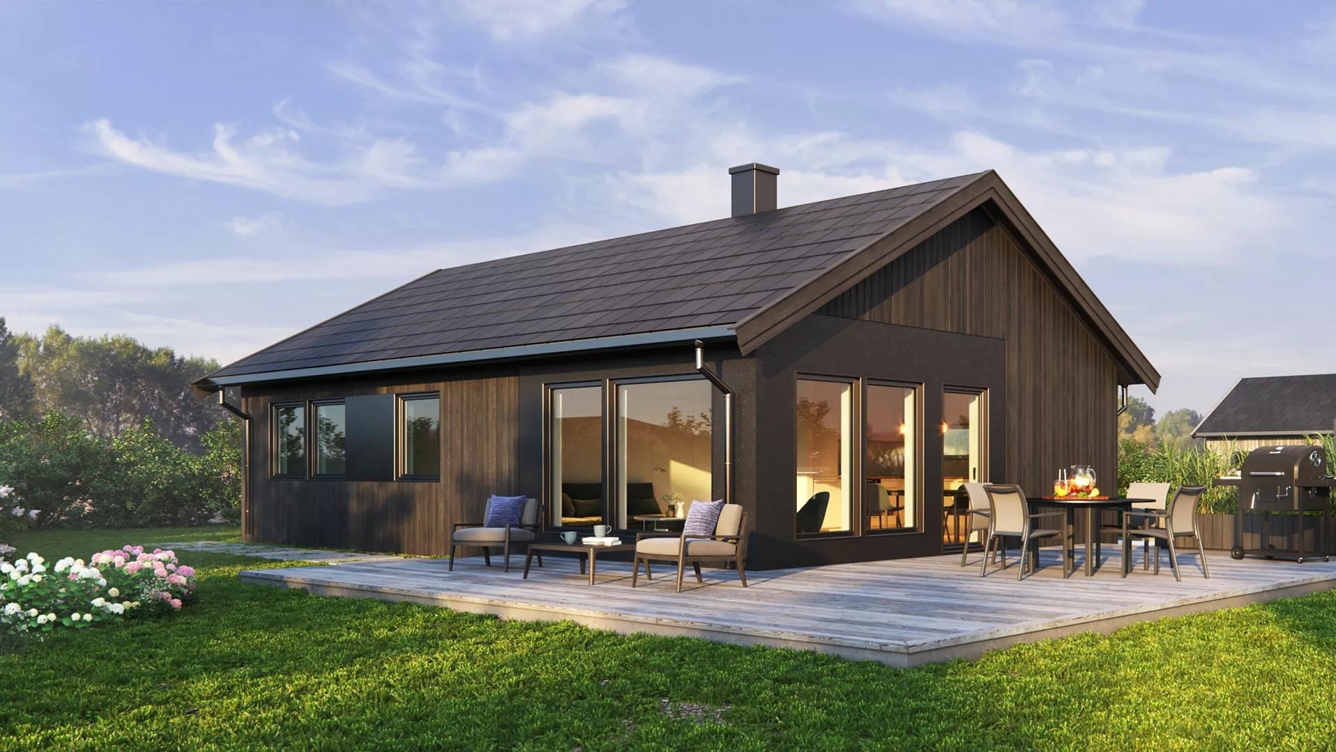 Nowoczesny dom szkieletowy z ciemną elewacją i dużymi oknami, otoczony zielonym trawnikiem i kwiatami, z tarasem i meblami ogrodowymi.