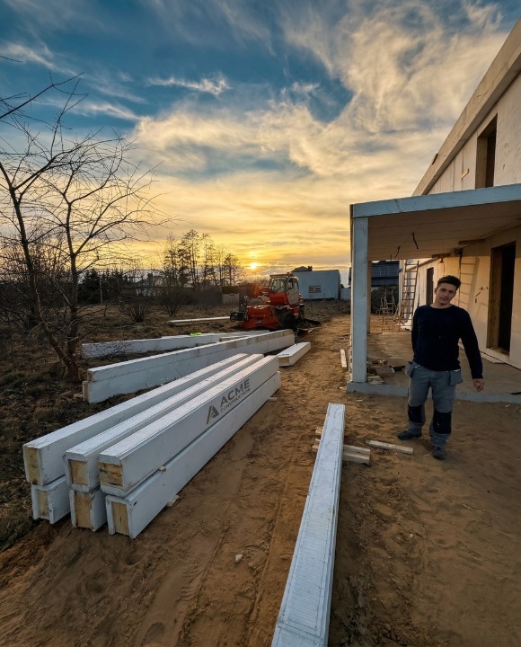 Budowa domu szkieletowego w trakcie: widoczne panele z logo ACME Construction, koparka i pracownik na tle zachodzącego słońca. Konstrukcja w stanie surowym otwartym.