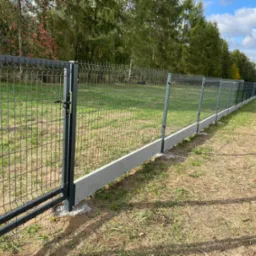 Długie, ciemne ogrodzenie z siatki panelowej z betonową podmurówką na nierównym terenie z widoczną bramą, zalesione tło i błękitne niebo z chmurami.