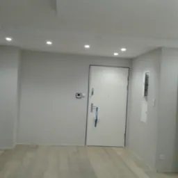 Wnętrze wykończonego pokoju z jasnymi, wyszpachlowanymi ścianami, drewnopodobną podłogą i białymi drzwiami wejściowymi z niebieską taśmą zabezpieczającą krawędzie ościeżnicy, oświetlonego...