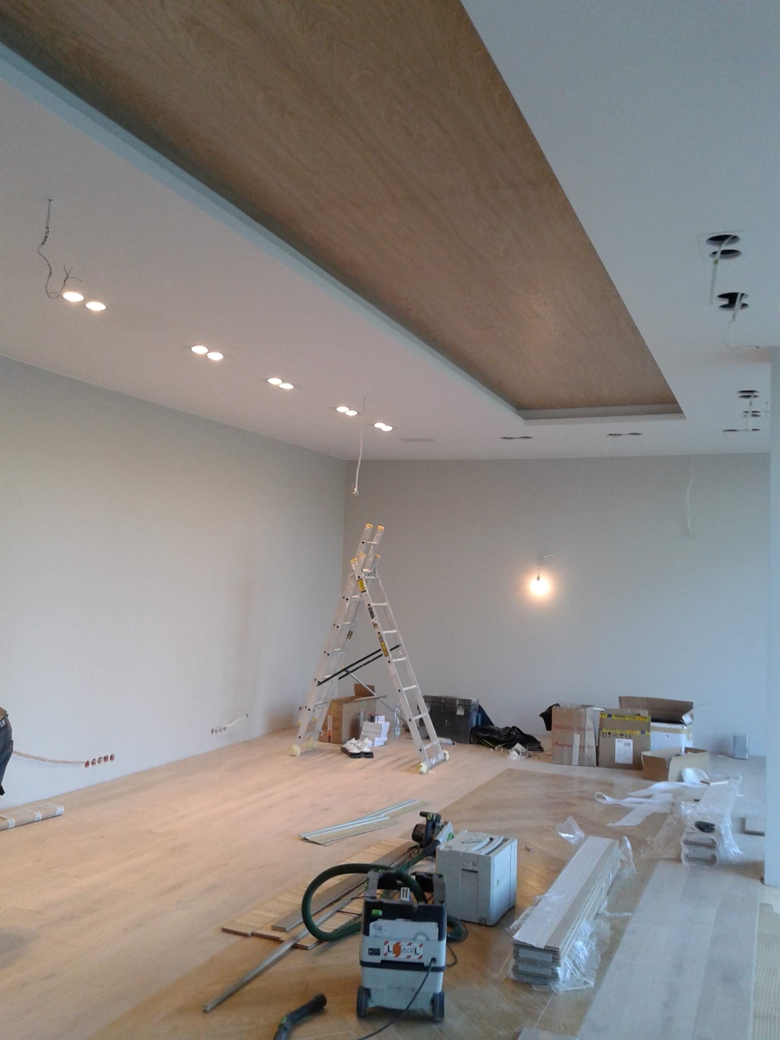 Wnętrze w trakcie prac wykończeniowych: drewniana wstawka w suficie, drabina, sprzęt budowlany i materiały rozłożone na podłodze.
