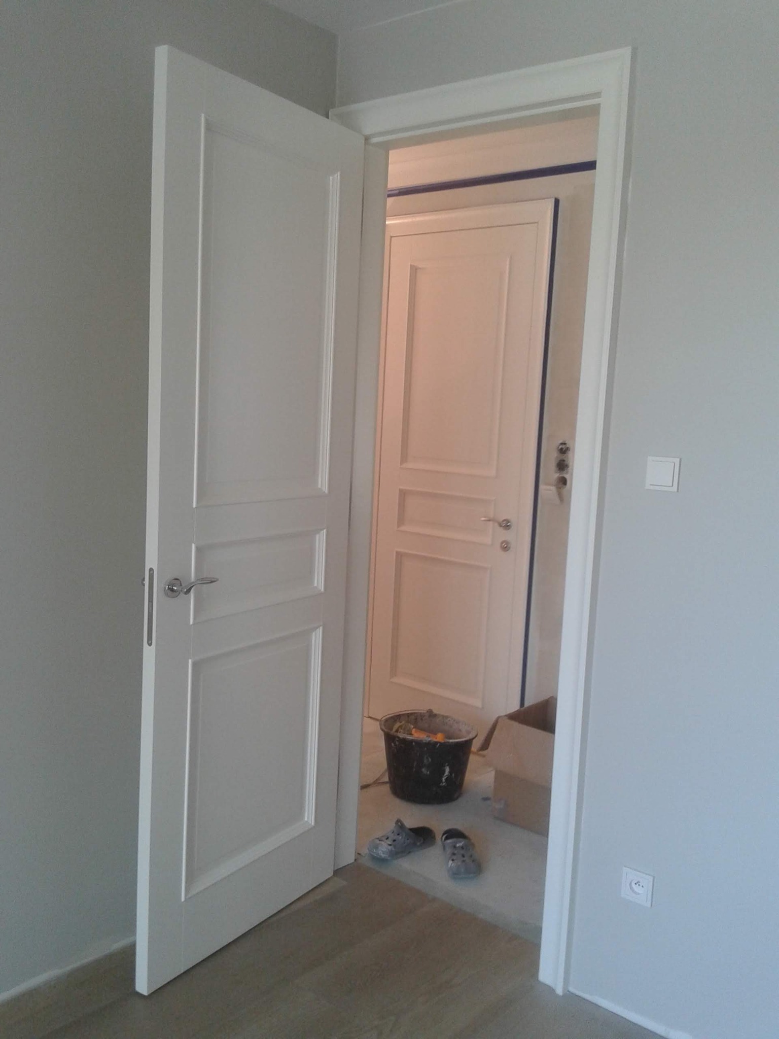 Otwarta, biała, płycinowa drzwi wewnętrzne z chromowaną klamką, widok na drugie drzwi zabezpieczone taśmą malarską, wiadro z narzędziami i robocze obuwie na podłodze.