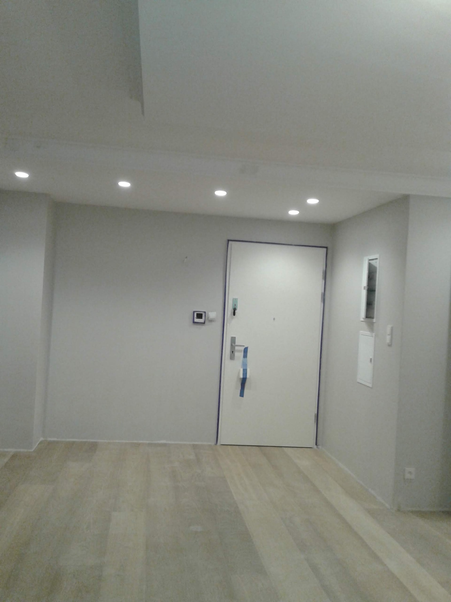 Wnętrze wykończonego pokoju z jasnymi, wyszpachlowanymi ścianami, drewnopodobną podłogą i białymi drzwiami wejściowymi z niebieską taśmą zabezpieczającą krawędzie ościeżnicy, oświetlonego...