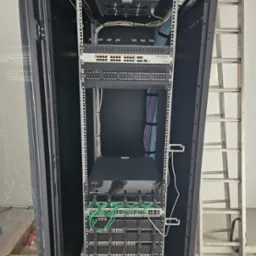 Otwarta szafa rack z zamontowanym sprzętem sieciowym, widoczne kable uporządkowane w wiązki, drabina oparta o ścianę w tle.