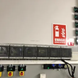 Panel sterowania systemem detekcji gazów z kilkoma centralami, przyciski awaryjne i czujniki, obok tablica informująca o gaśnicy do metali.
