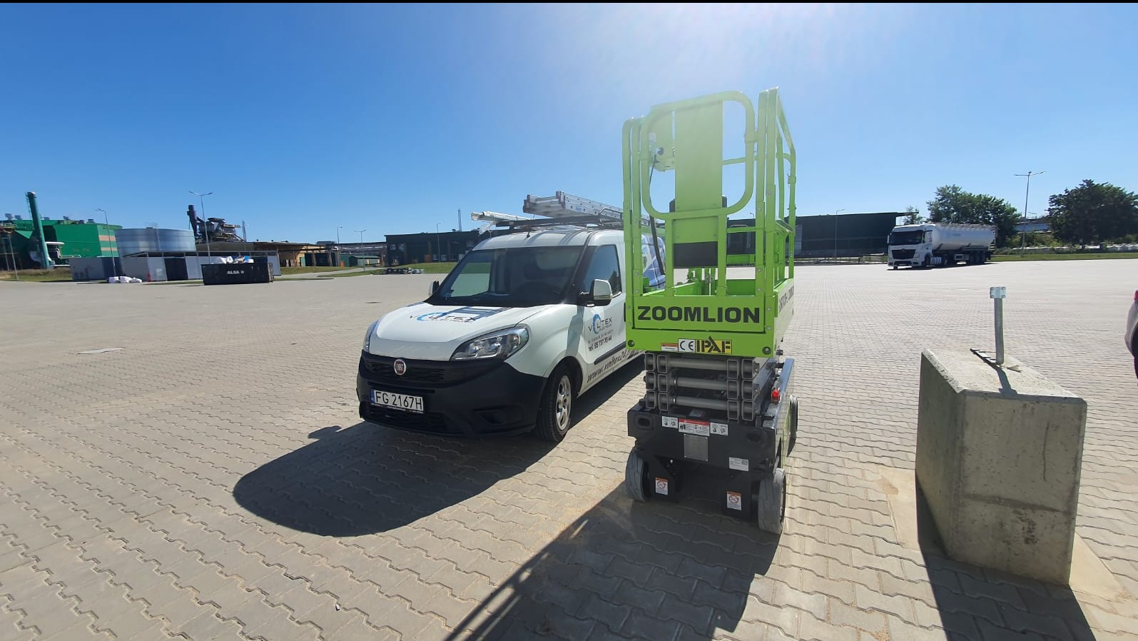 Biały samochód dostawczy z logo firmy Voltex oraz podnośnik koszowy Zoomlion na placu z kostki brukowej w słoneczny dzień.