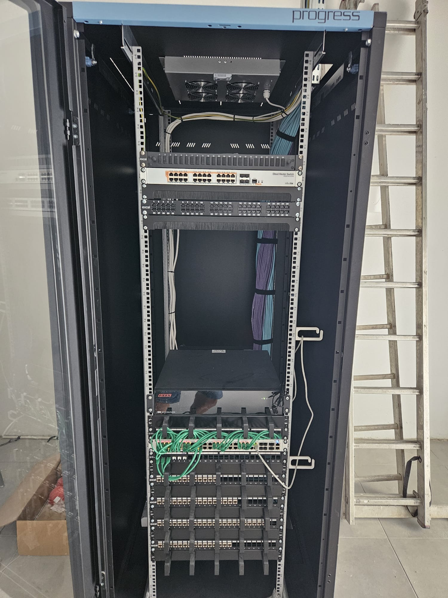 Otwarta szafa rack z zamontowanym sprzętem sieciowym, widoczne kable uporządkowane w wiązki, drabina oparta o ścianę w tle.