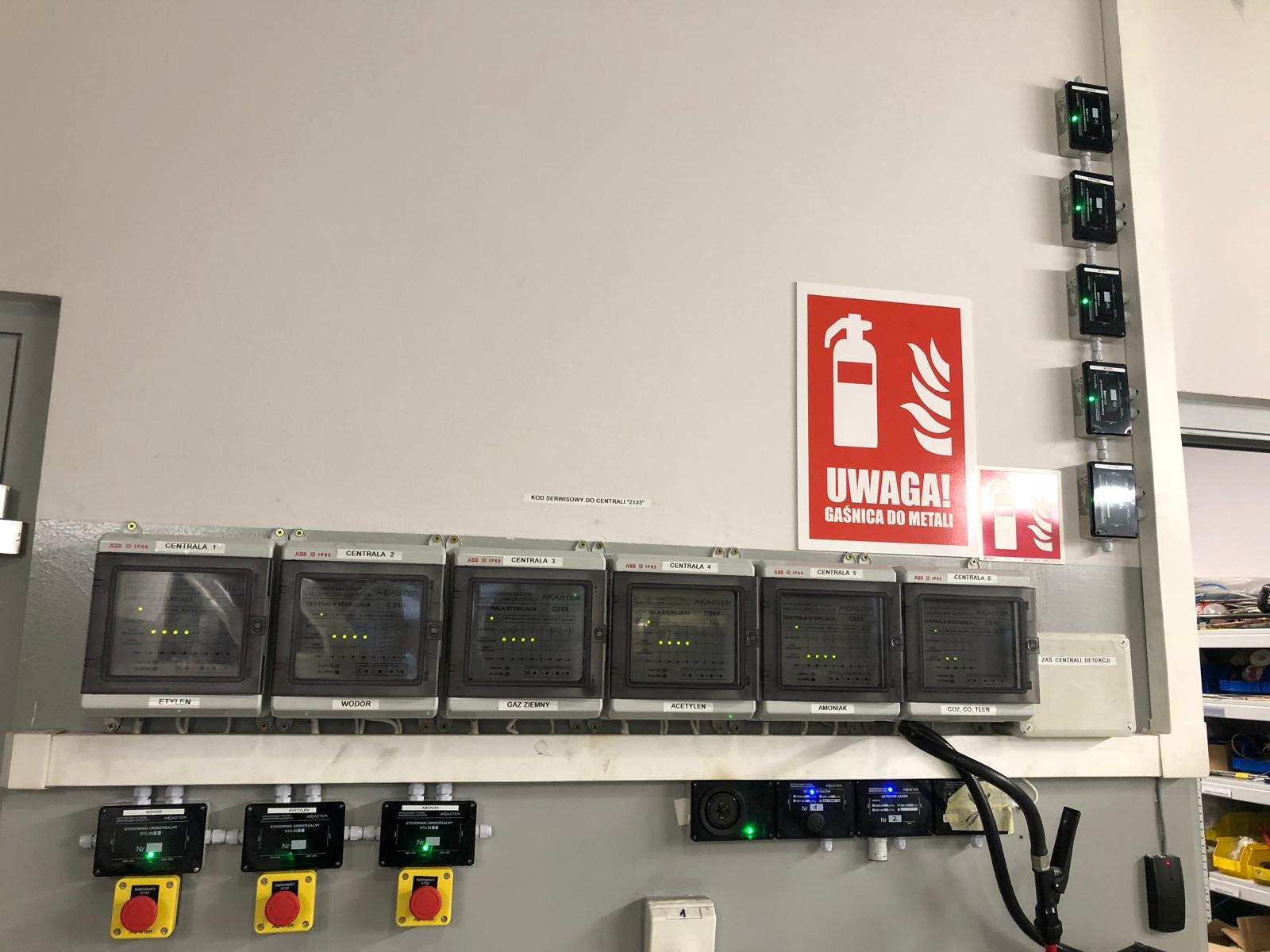 Panel sterowania systemem detekcji gazów z kilkoma centralami, przyciski awaryjne i czujniki, obok tablica informująca o gaśnicy do metali.