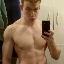 Młody mężczyzna robi selfie, prezentując swoją muskulaturę; widoczne napięte mięśnie ramion i klatki piersiowej na tle łazienki z ręcznikiem i szafką.