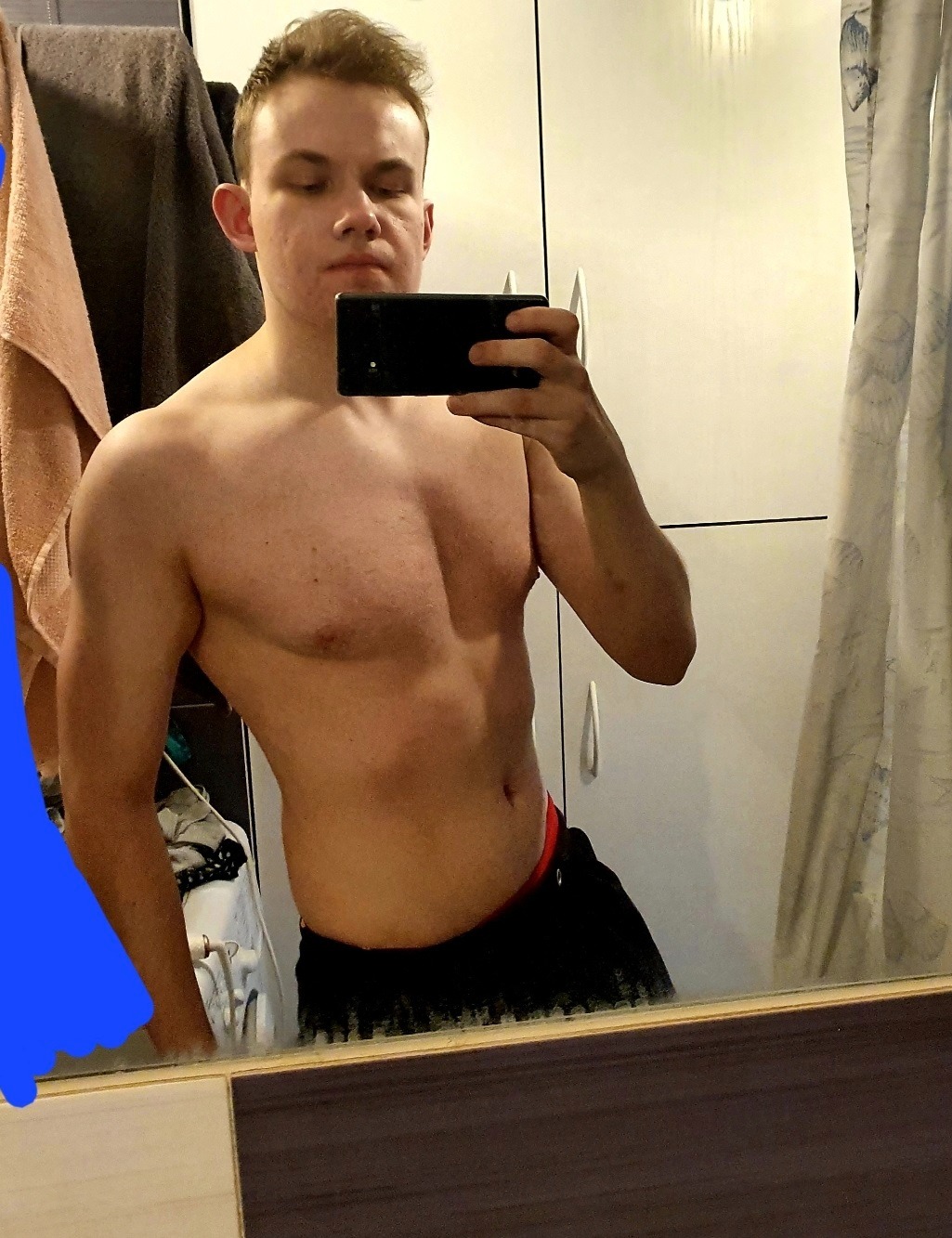 Młody mężczyzna robi selfie, eksponując umięśnioną klatkę piersiową i biceps, widoczne w odbiciu lustrzanym łazienki.