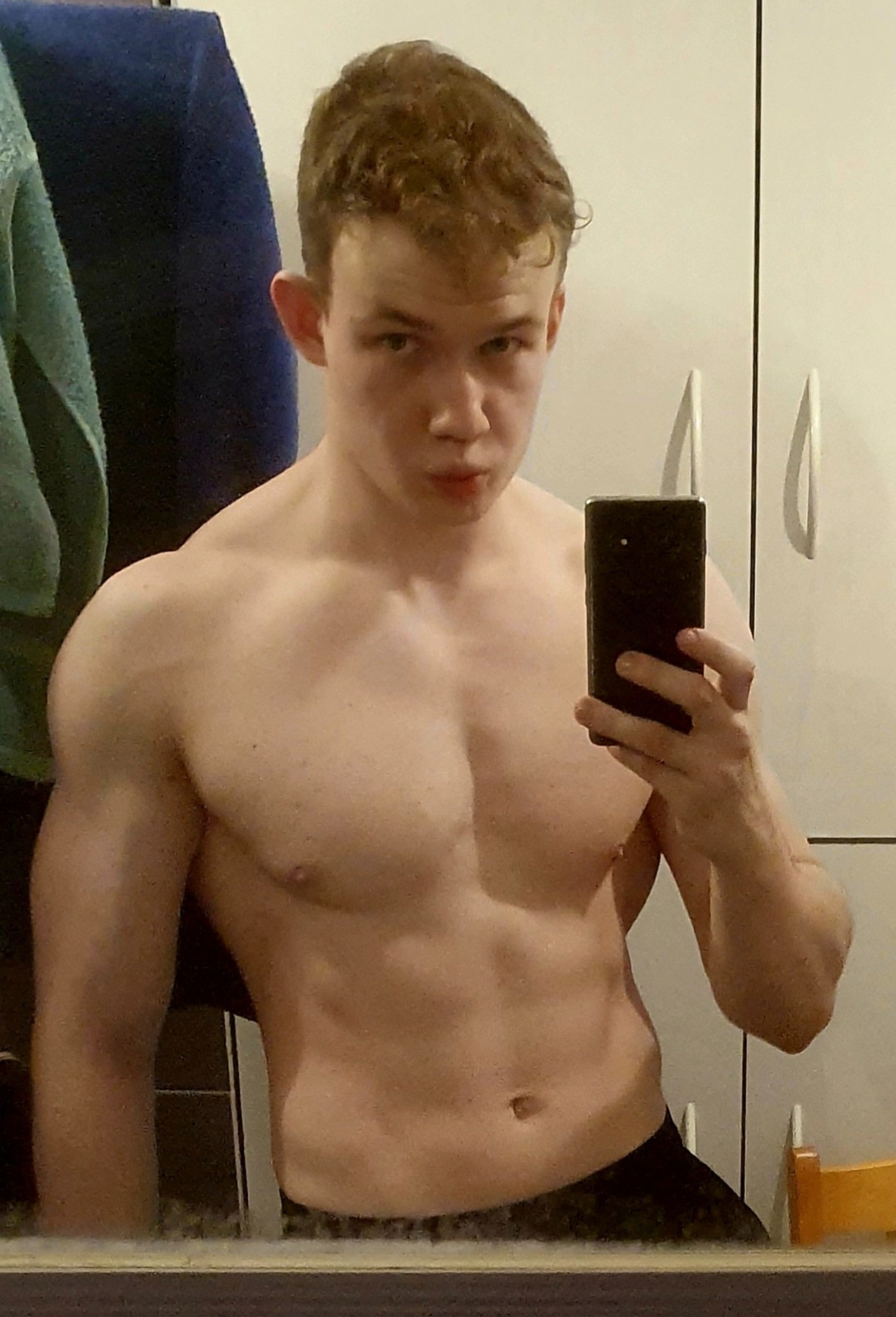 Młody mężczyzna robi selfie, prezentując swoją muskulaturę; widoczne napięte mięśnie ramion i klatki piersiowej na tle łazienki z ręcznikiem i szafką.