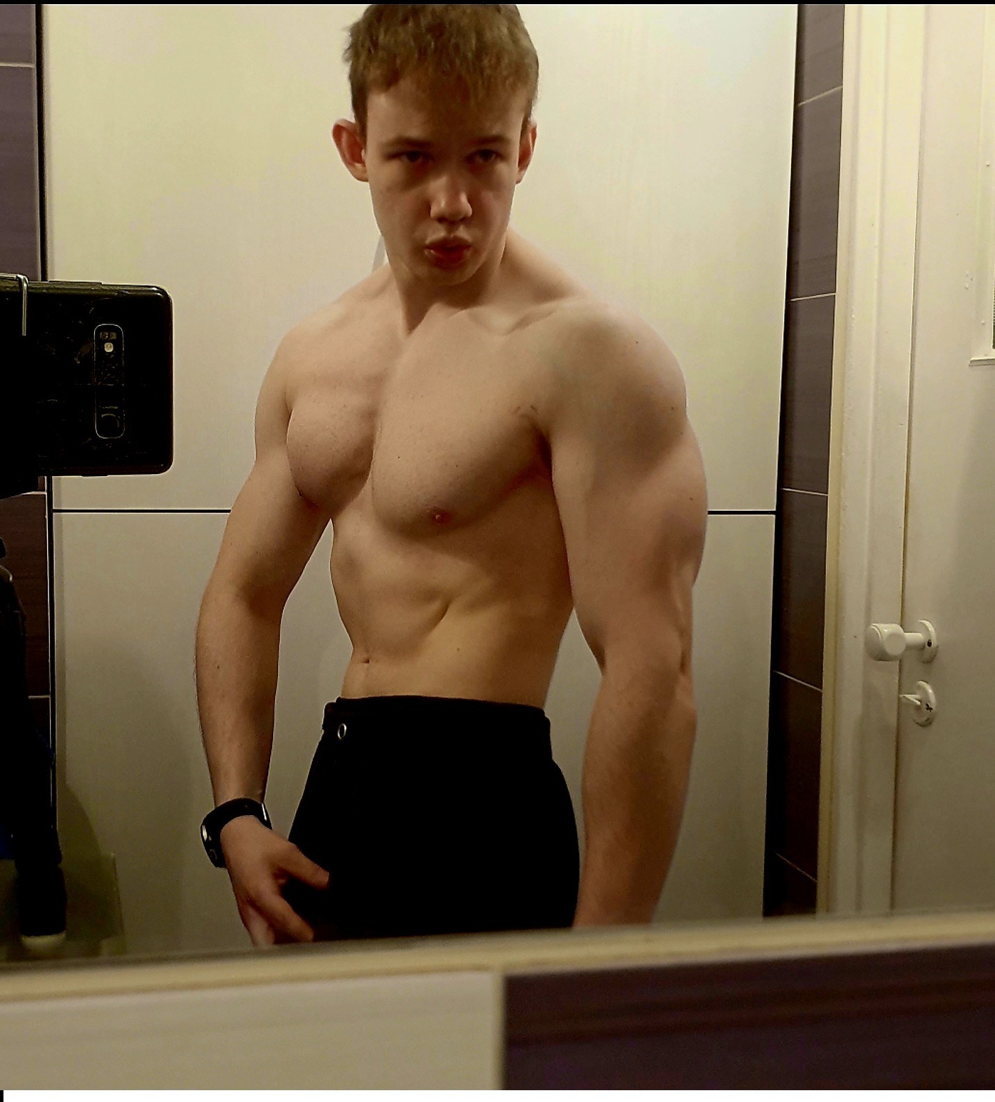 Mężczyzna prezentuje swoją muskularną sylwetkę, widoczne mięśnie klatki piersiowej, brzucha i ramion, pozując w łazience przed lustrem, z telefonem widocznym po lewej stronie kadru.