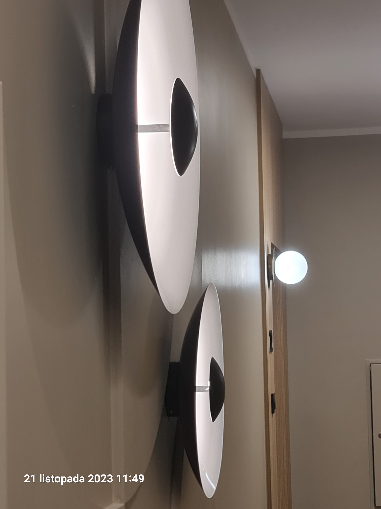 Dwa nowoczesne, okrągłe kinkiety ścienne z czarnymi elementami centralnymi, oświetlające beżową ścianę, obok pojedynczej lampy kulistej i drewnianej listwy ściennej.